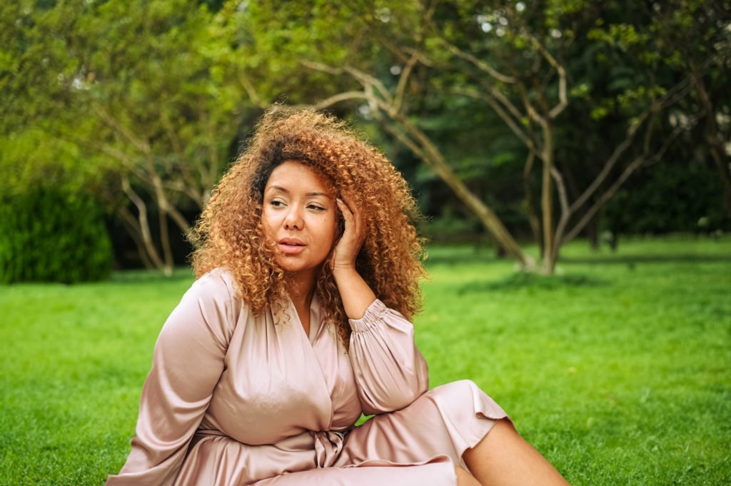Close up portrait of Young beautiful African American woman in summer public green park. The gorgeous plus size woman is wearing silk pink dress while sitting on grass and looking in the distance.