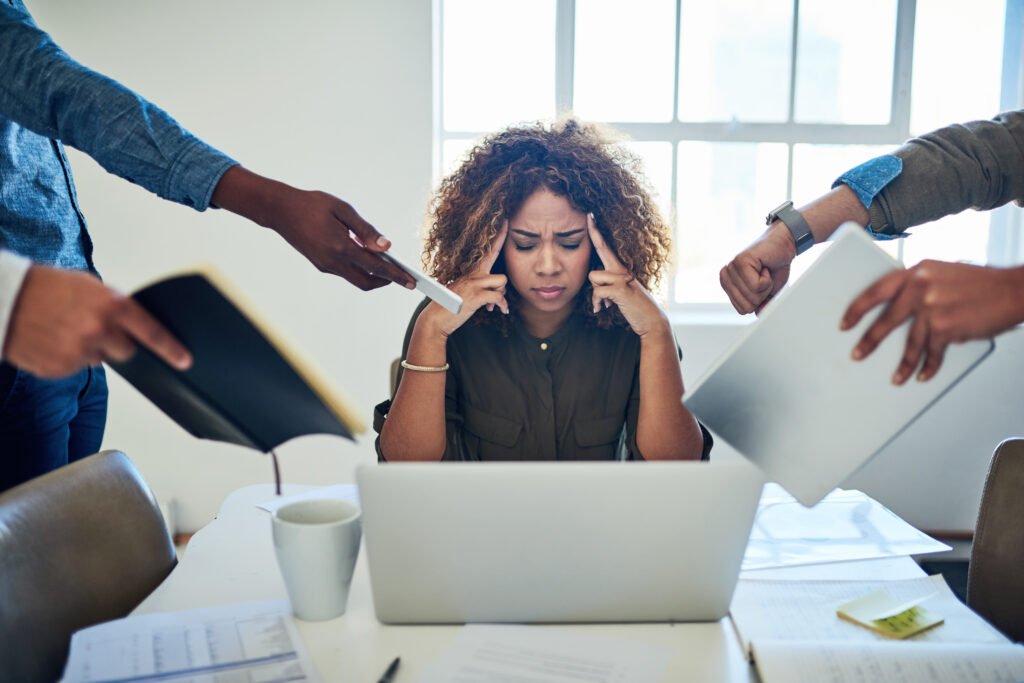 A Black woman with natural hair working at her laptop who is overwhelmed by a variety of requests.